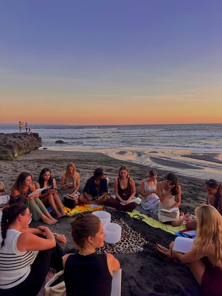 Groep mensen zittend in een cirkel op het strand bij zonsondergang met golven en rotsachtige kustlijn op de achtergrond