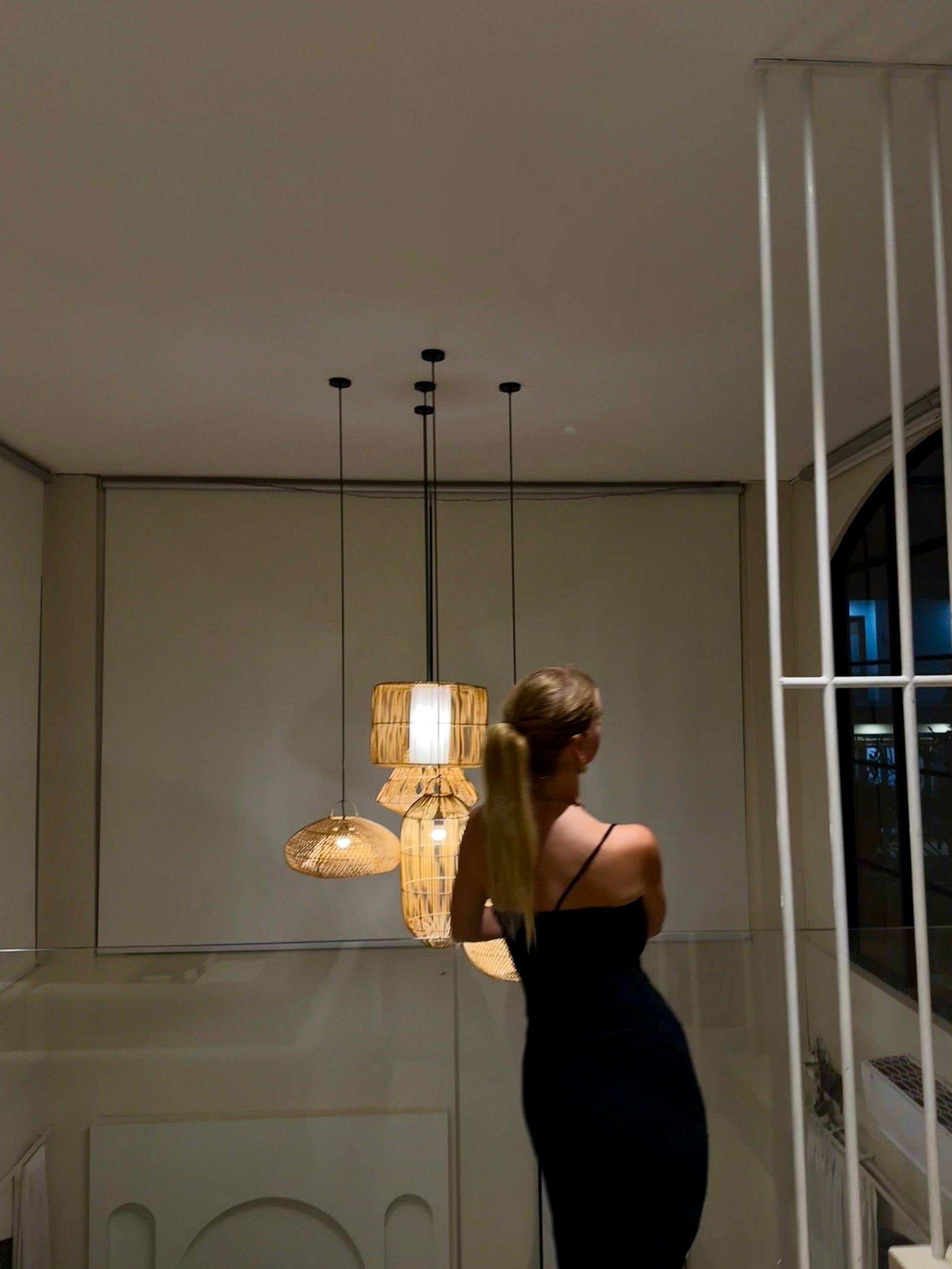 Vrouw in een zwarte jurk staand onder hangende lampen met natuurlijke houten kappen in een modern interieur