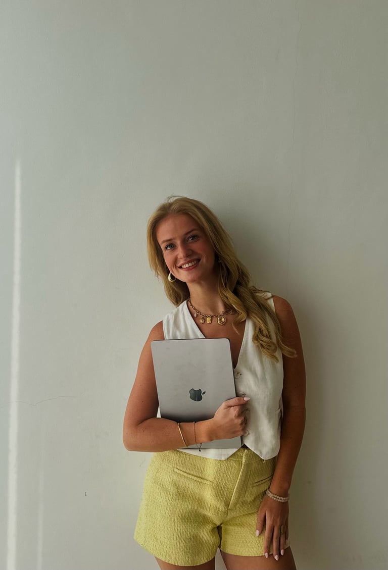Young woman smiling at camera holding a silver laptop, wearing a white sleeveless top and yellow shorts against a white wall