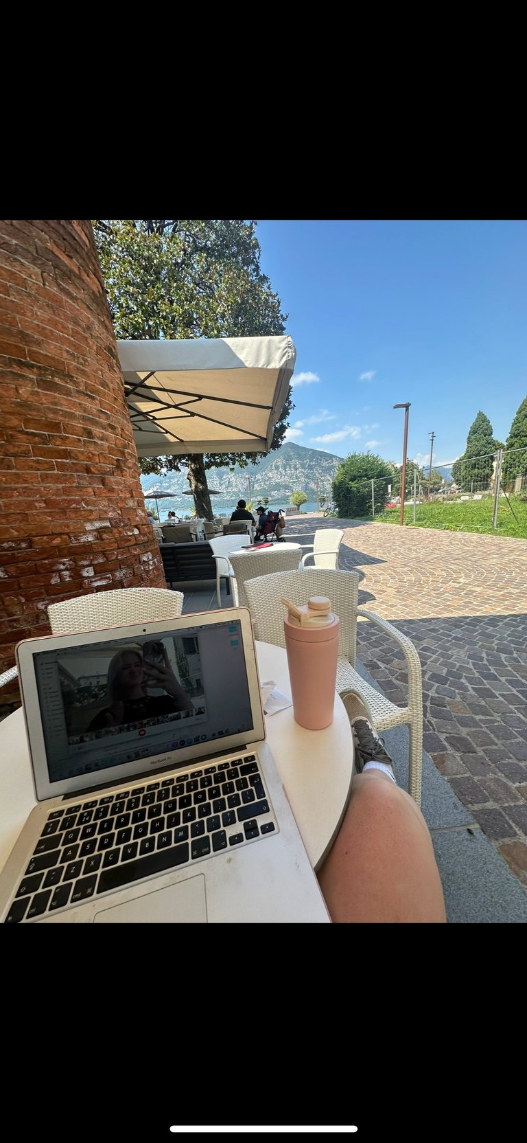 Person working on laptop at outdoor waterfront cafe with umbrellas and scenic mountain lake view