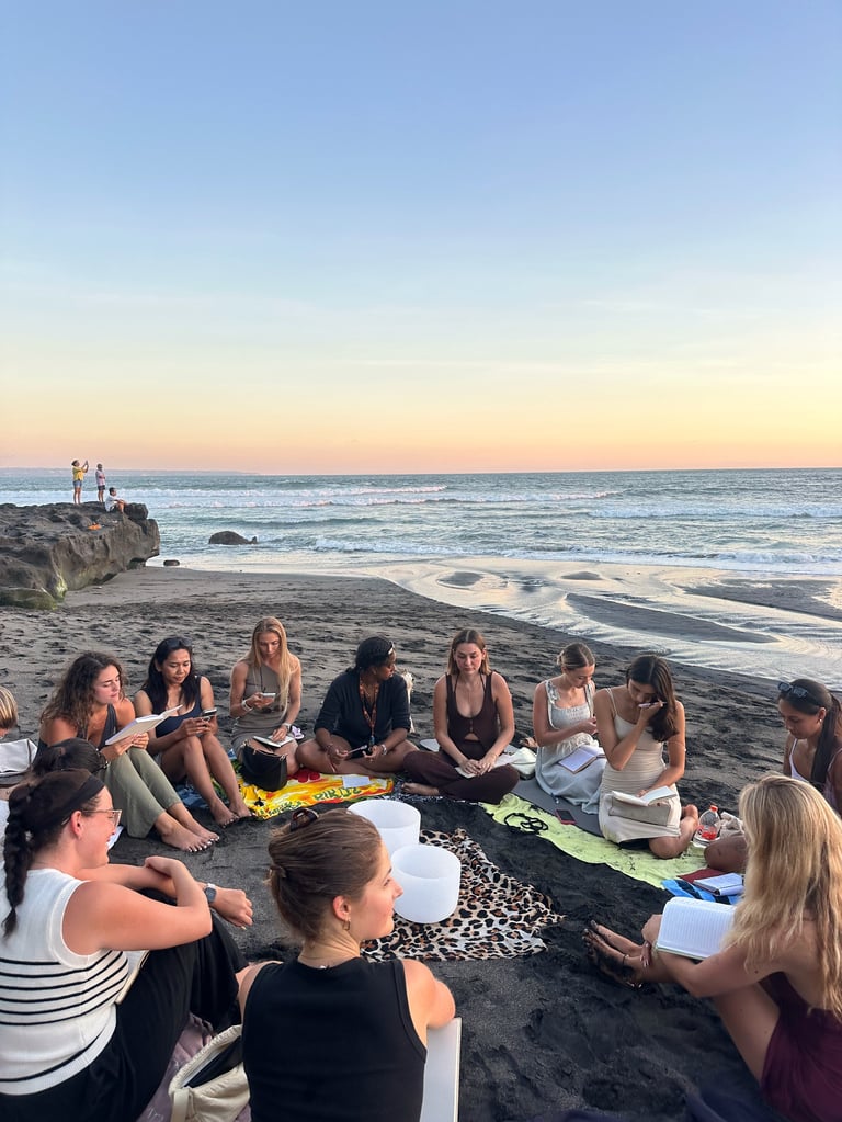 Group of people sitting in circle on beach at sunset with ocean waves and rocks in background