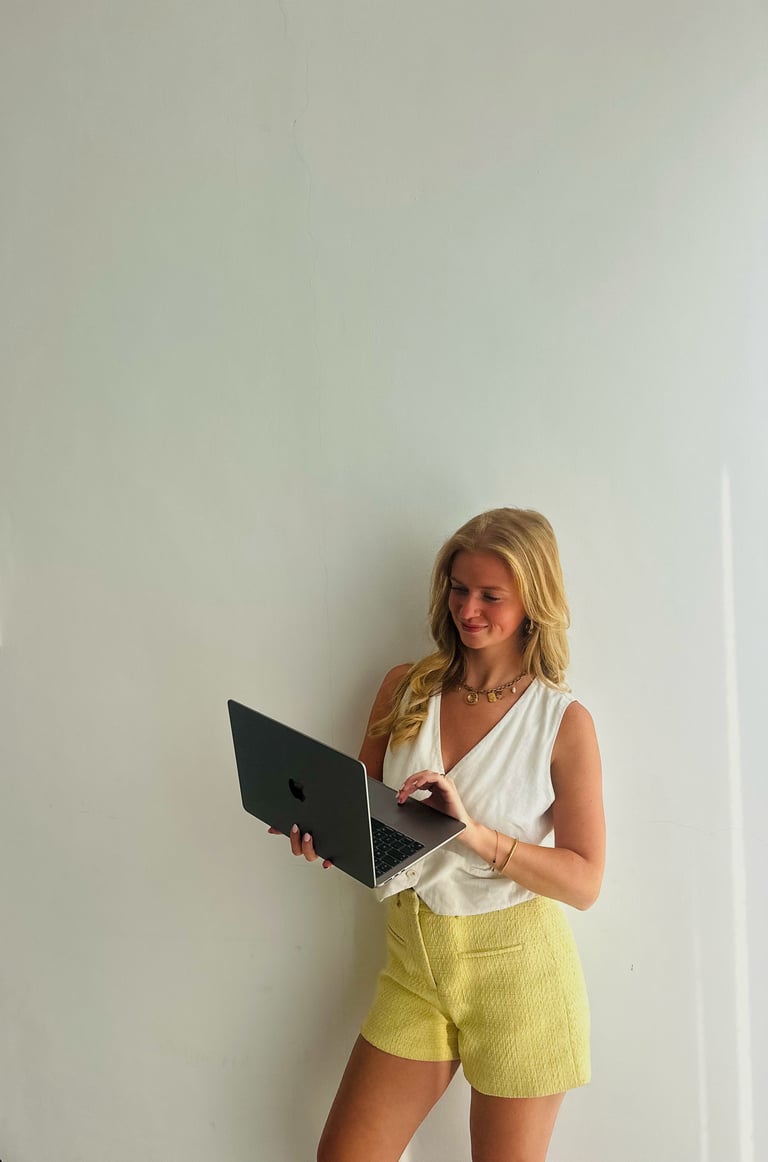 Vrouw met blond haar in een witte top en gele short, kijkend naar een laptop tegen een witte muur