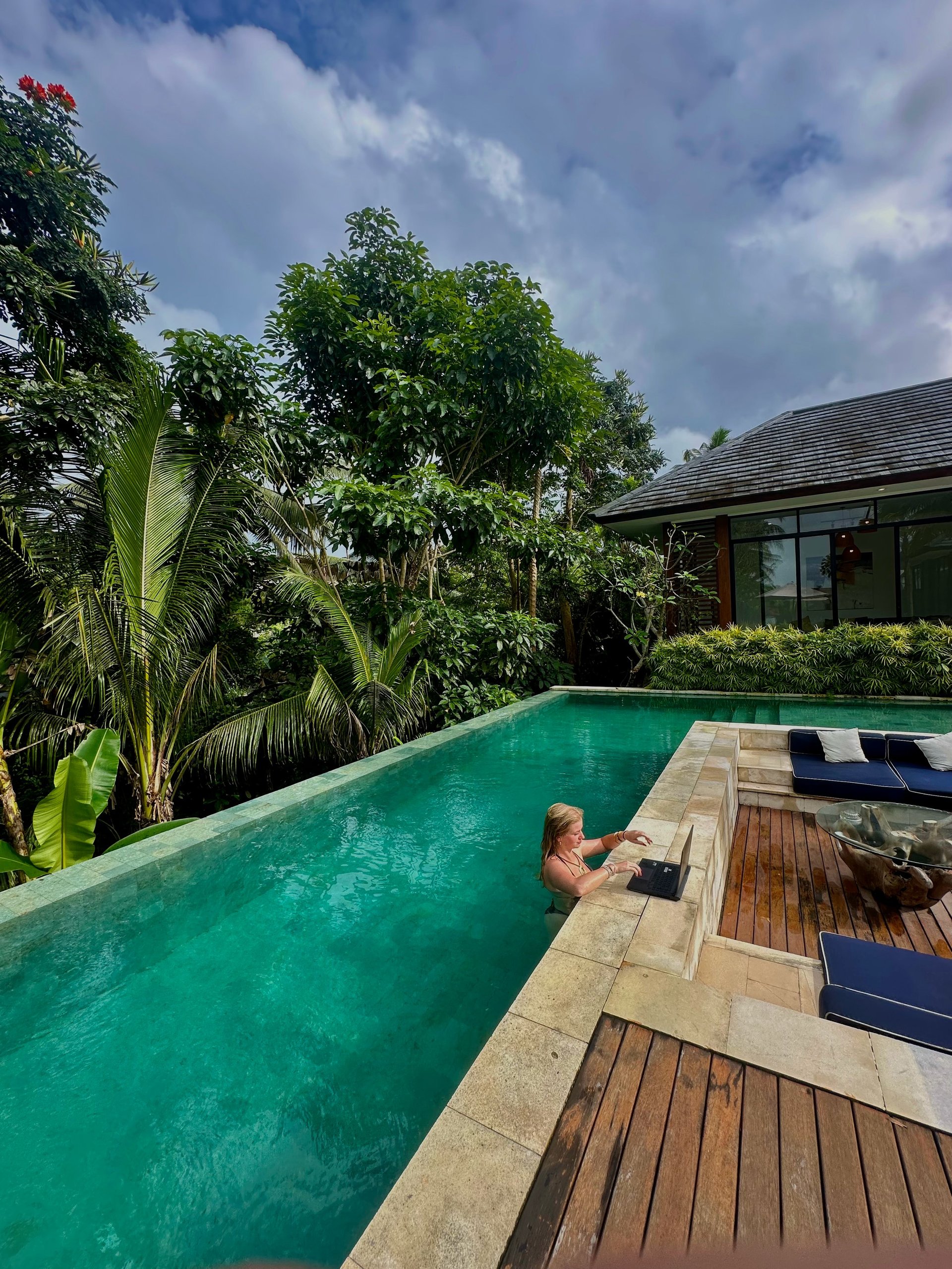 Woman swimming in tropical resort pool with wooden deck, loungers, and lush green vegetation under blue sky