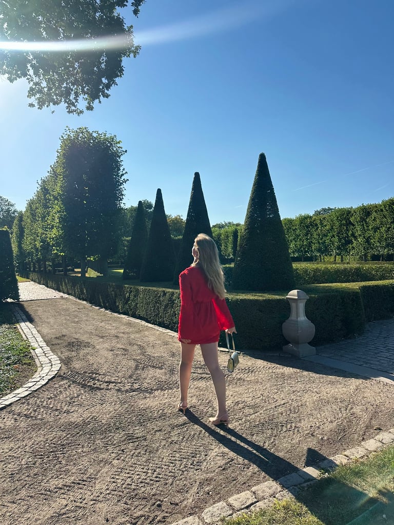 Vrouw in een rode jas staand op een tuinpad geflankeerd met hoge conisch gesnoeide struiken onder een blauwe hemel