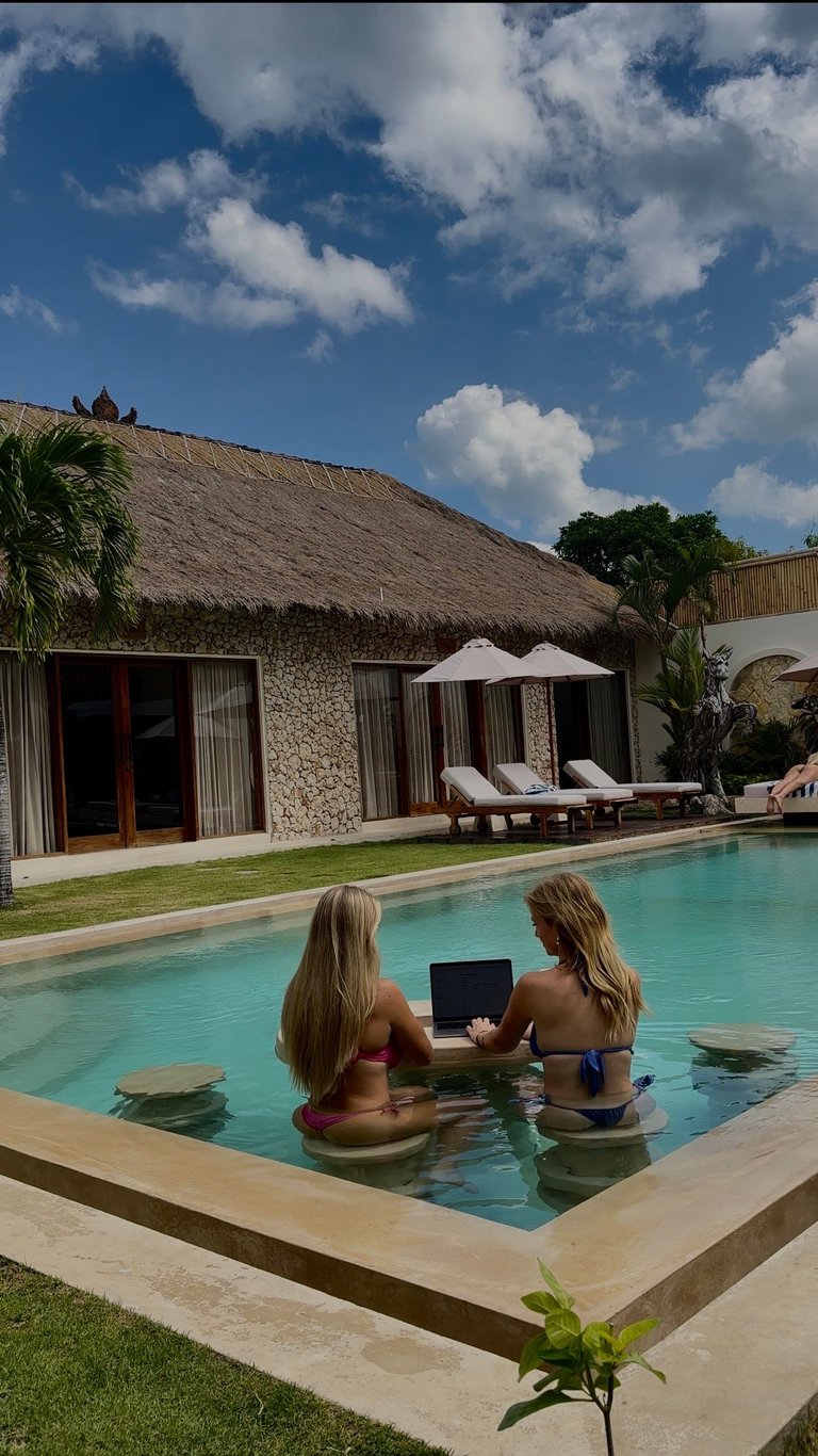 Twee vrouwen in een turquoise zwembad met laptops voor een huis met rieten dak, palmbomen en witte parasols